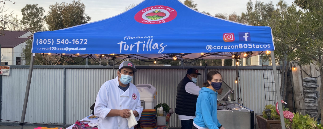 Corazon 805 Tacos operates an outdoor kitchen under a blue pop-up tent branded with their logo.