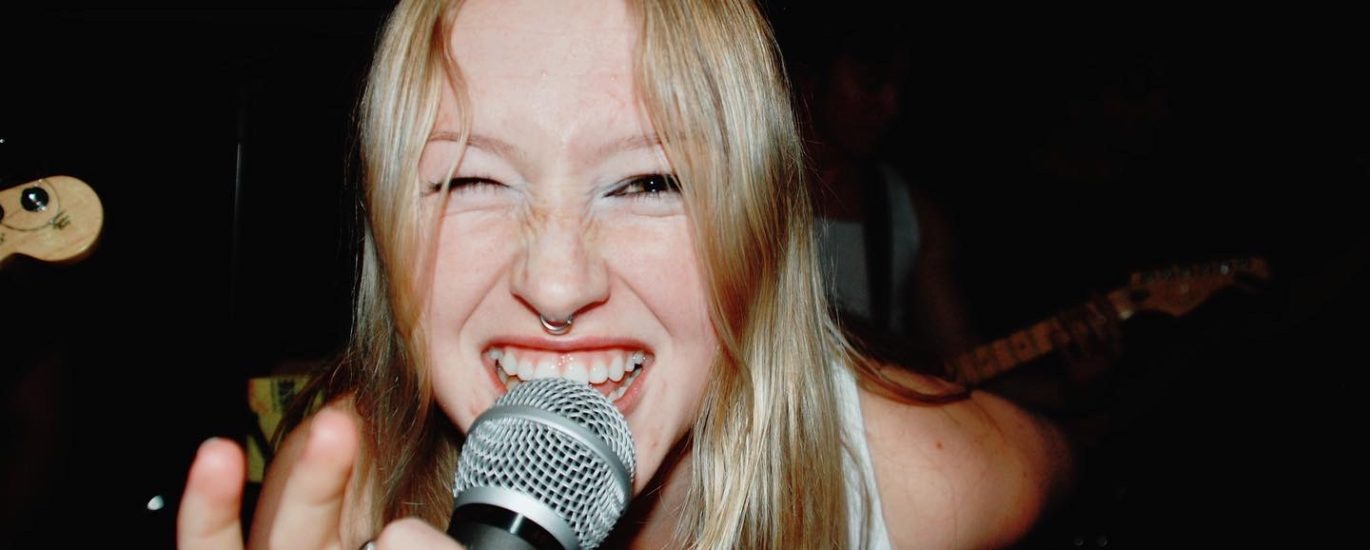 Skyllar Evans leaning forward into the camera, holding a microphone and smiling.