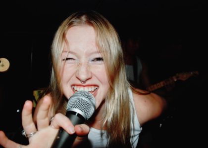 Skyllar Evans leaning forward into the camera, holding a microphone and smiling.