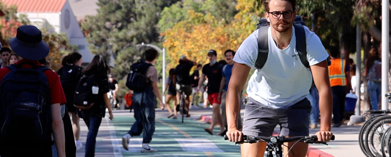 A man rides a bike in a bike lane through Cal Poly's campus. Pedestrians stroll in the background.