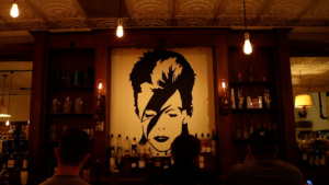 Three people are sitting at Highwater’s bar. We can see the backs of their heads. An image of David Bowie is pained on the wall behind the bar. 